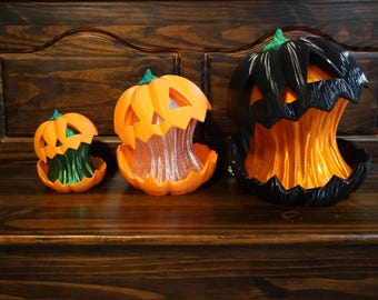May include: Three jack-o'-lantern decorations in varying sizes. The smallest is orange with a green interior, the middle is orange with a clear interior, and the largest is black with an orange interior. Each features a carved face design.