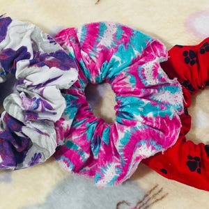 May include: Three fabric hair scrunchies arranged in a row. The first is white with a purple floral pattern. The second features a vibrant tie-dye design in shades of hot pink and bright turquoise. The third is red with a black paw print pattern.