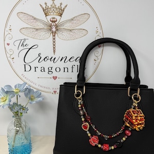 May include: A black structured handbag featuring a decorative double-strand beaded charm attached to the gold-tone hardware. The charm includes red heart-shaped beads, floral accents, and a miniature stack of red books with gold-tone detailing.