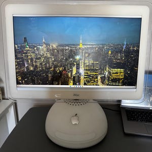 Puede incluir: Un ordenador Apple iMac blanco con un marco de plástico transparente que rodea la pantalla. La pantalla muestra un paisaje urbano nocturno de Nueva York con rascacielos iluminados. La base es una cúpula blanca redondeada con un logotipo de Apple plateado.