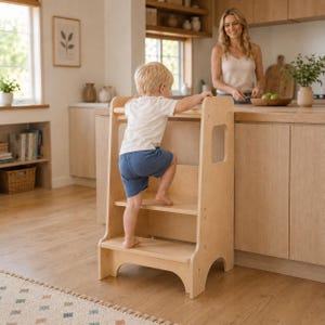 May include: A light-colored wooden learning tower with a two-step design and a safety rail at the top. The structure features smooth, unfinished wood surfaces, side cutouts for handles, and a stable base, providing a platform for reaching countertops.