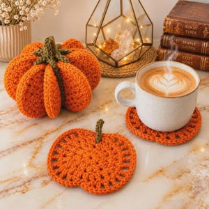 Puede incluir: Una calabaza naranja tejida a ganchillo con un tallo verde, un posavasos plano a juego con forma de calabaza y una taza de cerámica blanca llena de café con arte latte, dispuestos sobre una superficie de mármol con libros y una lámpara.