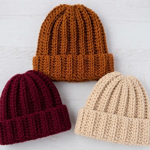 May include: Three knit beanies featuring a thick ribbed texture and a folded cuff. The hats are arranged in a triangular formation, with one beanie in a deep rust orange color, one in a dark burgundy red, and one in a light cream beige color.