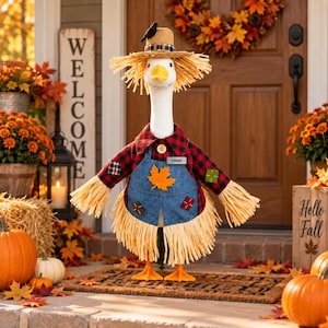 May include: A decorative goose figurine wearing a straw hat with a black bird, a red and black plaid shirt, and a blue denim overall dress featuring a yellow maple leaf patch and a name tag that reads EMMA. The goose has orange feet and straw trim.