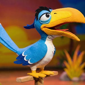 Puede incluir: Una marioneta de pájaro hecha de fieltro con un pico amarillo grande, plumaje azul y blanco, y plumas negras en la cabeza. El pájaro tiene el pecho blanco, una cola larga azul con detalles blancos y patas amarillas sobre un soporte de madera.