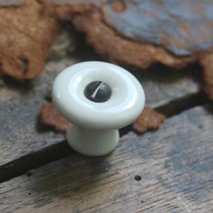 May include: A small, round, white ceramic knob with a smooth, glossy finish. The center of the knob features a dark, circular inset with a single flat-head screw visible in the middle, securing the piece to the surface below.