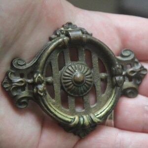 May include: Two antique-style brass drawer pulls with a circular ring handle and a decorative backplate featuring vertical slats and a central rosette. The metal hardware has a dark, aged patina with highlights on the raised edges of the rings.