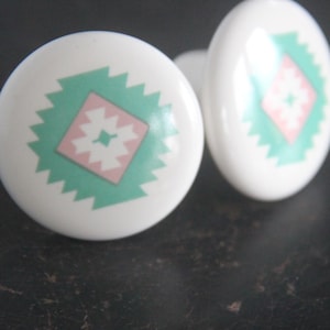 May include: Two white ceramic cabinet knobs with a geometric design. Each knob features a central pink diamond shape surrounded by a green zigzag pattern. The knobs are set against a dark background.