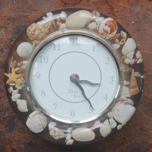 May include: A round clock with a white face and black hands, surrounded by seashells and a starfish. The clock is set in a clear resin and has a silver-colored metal frame. The clock is on a brown, textured surface.
