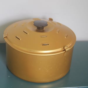 May include: A round, metallic gold-colored canister with a matching lid. The lid features a dark brown, knob-style handle at the center and several small, rectangular ventilation slits arranged in a circular pattern around the top surface.