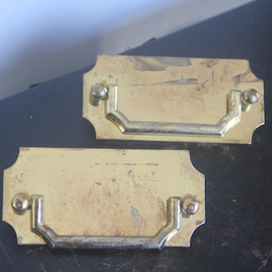 May include: Two vintage brass drawer pulls with a rectangular backplate and a curved handle. The pulls have a tarnished, aged appearance, with visible wear and tear. They are mounted with small, round screws.
