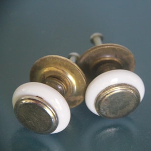 May include: Two cabinet knobs featuring a smooth white ceramic ring surrounding a circular metallic center piece. Each knob is attached to a brass-toned metal base with a visible mounting screw extending from the back of the hardware.