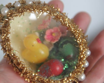 May include: A gold, pearl, and glitter Easter egg ornament with a yellow rubber ducky, red and pink flowers, and green foliage inside.