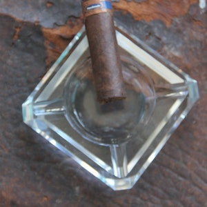 May include: A clear glass ashtray with a square shape and rounded corners. A brown cigar is resting in the ashtray.