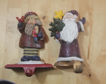 May include: Two Christmas stocking holders shaped like Santa Claus figures. One Santa holds a bird and a nutcracker, the other holds a Christmas tree and a star. The figures are painted in shades of red, purple, and green.