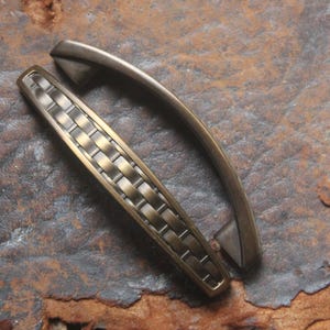 May include: Two metal cabinet pulls with an antique brass finish. One pull features a smooth, arched design, while the other displays a textured, woven-style pattern along its center. Both hardware pieces have a curved shape and metallic sheen.