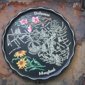 May include: A black, round tray featuring a map of Maryland and Delaware. The tray has a scalloped edge and is decorated with floral illustrations, including pink blossoms and yellow Black-Eyed Susans. The words "Delaware" and "Maryland" are displayed.