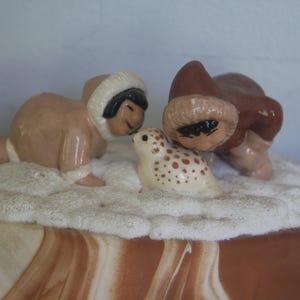 May include: A ceramic figurine set featuring two figures in brown hooded parkas with white fur trim, positioned on a textured white base. The figures are leaning toward a small, cream-colored seal pup with brown spots, all set on a marbled brown surface.