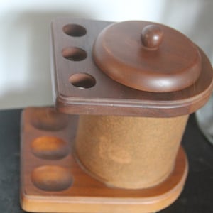 May include: A wooden pipe rack with a round lidded container. The rack has four slots for pipes and a round base with three indentations. The container is made of wood and has a light brown finish.