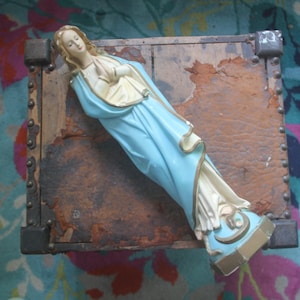 May include: A religious figurine of a woman with long hair, wearing a light blue and cream-colored robe with gold trim. The figure is depicted with hands clasped together in a prayer position, standing on a small, hexagonal base with gold accents.