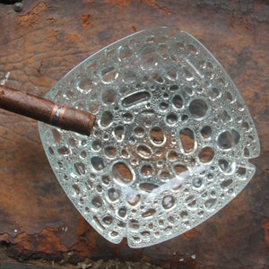 May include: A clear glass ashtray with a textured bubble design. A brown cigar rests on the edge of the ashtray.