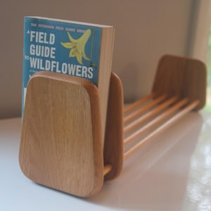 May include: A wooden book holder with a book titled "Field Guide to Wildflowers" is displayed. The book holder is made of light-colored wood and has a rectangular base with a series of wooden rods. The book has a blue cover with white text and a yellow flower illustration.