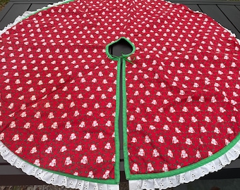 May include: Red Christmas tree skirt with a repeating pattern of white snowmen and green holly. The skirt has a green border and a white eyelet lace edge. It is designed to wrap around a tree trunk.