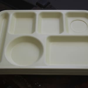 May include: A stack of off-white, compartmentalized trays. Each tray has six sections of varying shapes and sizes, including a circular recess for a cup. The trays are made of a smooth, durable material.