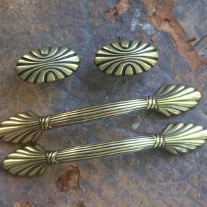 May include: Two oval-shaped cabinet knobs and two matching elongated drawer pulls, all crafted from a metallic brass-toned material. The knobs feature a radiating sunburst pattern, while the pulls have a ribbed center and fan-shaped ends.