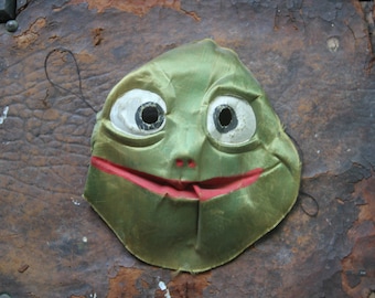 Puede incluir: Una máscara de tela verde con ojos blancos grandes y una boca roja sonriente. La máscara está colocada sobre una superficie de cuero marrón desgastado. La máscara tiene un diseño caprichoso y caricaturesco.