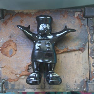 May include: A black ceramic chef figurine with outstretched arms, wearing a chef's hat and uniform. The figurine is placed on a weathered wooden surface with metal accents, suggesting a vintage or rustic aesthetic.