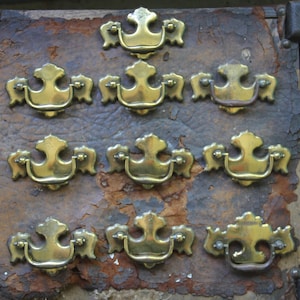 May include: Eight antique brass drawer pulls with ornate designs. The pulls are arranged in a triangular pattern on a distressed brown leather surface.