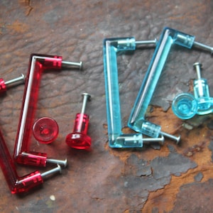 May include: Assortment of red and blue plastic cabinet knobs. The knobs are shaped like a simple rectangle with rounded corners. The knobs have a hole in the center for a screw.
