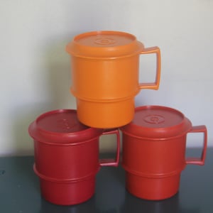 May include: Three vintage plastic mugs with lids. One is orange, and two are red. Each mug has a handle and a lid with a circular design. The mugs are stacked on top of each other.