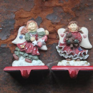 May include: Two Christmas stocking holders featuring angel figurines. The angels have red dresses, white wings, and hold a star and a teddy bear. The holders are red with a curved design, and the figurines are on a white cloud base.