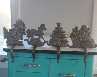May include: Four antique metal Christmas stocking holders. The set includes Santa Claus, a rocking horse, a Christmas tree, and Santa in a sleigh. Each holder has a hook for hanging stockings. The holders are silver in color.
