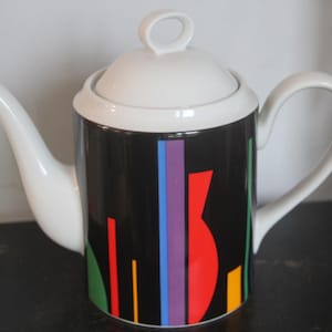 May include: White ceramic teapot with a black, red, green, blue, and purple geometric design. The teapot has a lid and a handle.