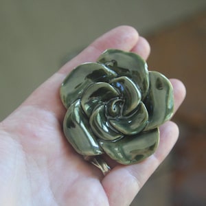 May include: A glossy, olive-green ceramic flower brooch with layered, curved petals. The surface of the ceramic piece has a reflective, smooth finish, showcasing a deep, earthy green color with subtle variations in tone across the sculpted petals.