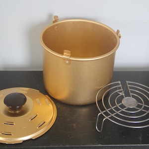 May include: A gold-colored, vintage-style cooking pot with a matching lid and a metal trivet. The pot has a rounded shape with a lid and a black knob. The trivet is made of silver-colored metal.
