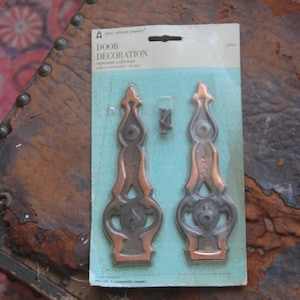 May include: Package of two decorative door hardware pieces. The pieces are made of metal and have a copper and black finish. The package includes screws for installation. The package is labeled "Door Decoration".