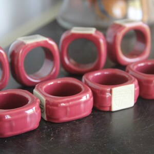 May include: A collection of eight square, burgundy napkin rings. Each ring has a square shape with rounded corners and a cream-colored accent on one side. The rings are arranged on a dark surface.
