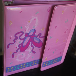 May include: Pink binder with a design of two pink ballet shoes with a white and blue swirl pattern. The binder has a blue, green, and yellow striped border.
