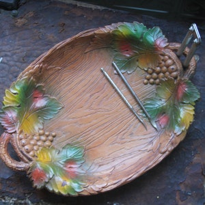 May include: A wooden serving tray with a grapevine design. The tray is painted with brown, green, and red colors. Two silver tools are placed on the tray.
