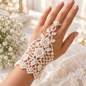 May include: An off-white crochet hand flower bracelet featuring an intricate lace pattern with floral motifs, small leaves, and tiny white pearls. The design includes a wrist cuff connected by a delicate chain of lace to a matching ring.