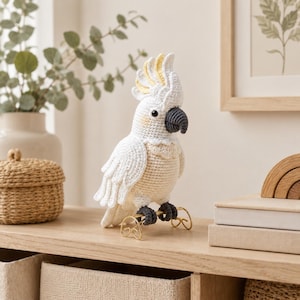 May include: A crocheted cockatoo figurine featuring white plumage, a yellow-tipped crest, a dark gray beak, and dark gray feet. The bird is perched on a small, gold-colored wire stand, showcasing detailed stitching throughout its body and wings.