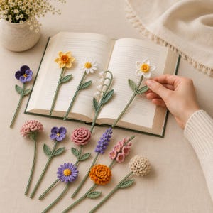May include: A collection of crocheted flower bookmarks featuring various species including a yellow daffodil, white daisy, purple pansy, pink rose, orange marigold, and lavender sprig, each with green stems and leaves, displayed with an open book.