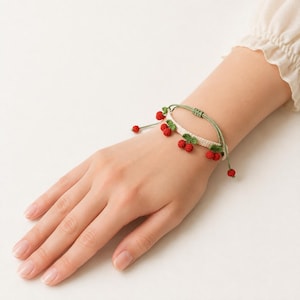 Puede incluir: Una pulsera tejida con una banda de color crema adornada con varios pares de pequeñas cerezas de ganchillo rojas y hojas verdes. La pulsera se asegura con un cordón verde fino y ajustable que se ata con detalles de cerezas colgantes.