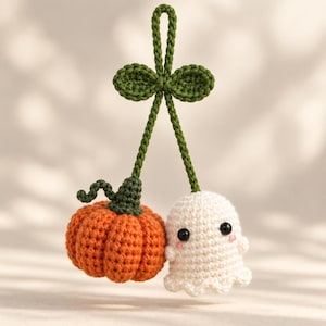 May include: A crochet hanging ornament featuring an orange pumpkin and a white ghost connected by green stems. The pumpkin has a dark green stem and the ghost has black bead eyes with small pink cheeks. A green loop at the top allows for hanging.