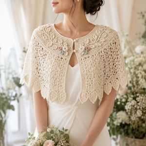 Puede incluir: Un bolero de ganchillo color crema con un intrincado patrón de hojas a lo largo del escote y el dobladillo, delicados detalles florales en tonos pastel suaves y dos pequeños botones blancos nacarados en el cierre frontal sobre un vestido blanco.