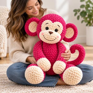 May include: A large, plush, crocheted monkey toy featuring a vibrant magenta body and limbs with contrasting cream-coloured hands, feet, ears, and face. The monkey has black safety eyes, a brown embroidered nose, a stitched smile, and a curled tail.